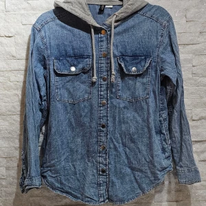 Jeansskjorta med grå huva från H&M Divided - Säljer en snygg jeansskjorta från H&M Divided i ljusblå denim med grå huva och dragsko. Skjortan har två bröstfickor med knappar och långa ärmar. Perfekt för en avslappnad och cool stil. Passar s/m oxå! Se bilder då jag är en s/m