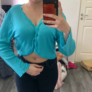 Turkos croppad kofta från Street One - Säljer en turkos croppad kofta från Street One med v-ringning och spetsdetaljer längs halsringningen. Tröjan är ribbad och har långa ärmar samt knyt framtill. Perfekt för dig som vill ha en färgglad och trendig topp.