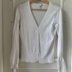 Vit kofta från H&M - Säljer en klassisk vit kofta från H&M i storlek M men passar S också. Koftan har långa ärmar, v-ringning och knäppning framtill med vita knappar. Ribbstickade muddar vid ärmslut och nederkant. Perfekt att slänga över en topp eller klänning.