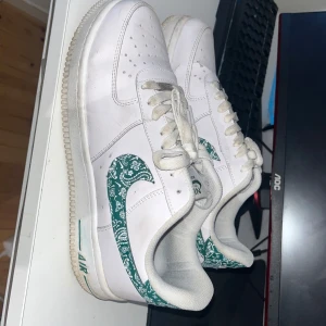 Nike Air Force 1 med grön paisley-detalj - Säljer ett par vita Nike Air Force 1 med unika gröna paisley-mönstrade detaljer på swoosh och häl. Klassisk siluett med snörning och perforerad tå för ventilation. Perfekta för dig som vill sticka ut med en personlig touch på dina sneakers. Passar även 43 och pris kan diskuteras 