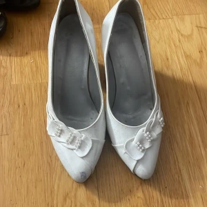 Vita pumps med klack och dekorativa rosetter - Säljer ett par vita pumps med klack och spetsig tå. Skorna har fina rosetter framtill dekorerade med pärlor och paljetter. Perfekta för dig som vill ha något elegant och klassiskt.
