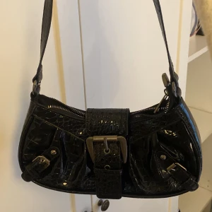 Svart axelväska med krokodilpräglat mönster - Säljer en svart axelväska i glansigt syntetmaterial med krokodilpräglat mönster. Medföljer matchande hjärtformat tillbehör och liten plånbok som går att sätta på. Oanvänd bara legat i garderoben. 