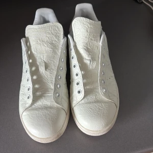 Vita Adidas Stan Smith sneakers - Säljer ett par klassiska vita Adidas Stan Smith sneakers i skinn. Skorna har en stilren design med präglad Adidas-logga på hälen och sidan. Perfekta för dig som gillar minimalistisk stil och vill ha ett par tidlösa sneakers.