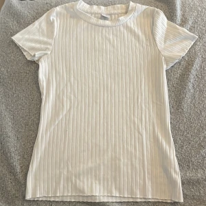 Vit ribbad t-shirt från Lindex - Säljer en vit ribbad t-shirt från Lindex i storlek 158/164. T-shirten har korta ärmar och rund halsringning. Perfekt basplagg med snygg ribbad struktur som passar till det mesta. 💕 Dock så står det ett namn på baksidan av storleks lappen men det är inget som syns💕