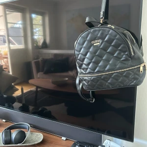 Svart quiltad ryggsäck från Guess - Säljer en svart quiltad ryggsäck från Guess med guldfärgade dragkedjor och logga framtill. Väskan har flera fack och justerbara axelremmar. Perfekt för dig som vill ha en snygg och praktisk accessoar till vardagen.