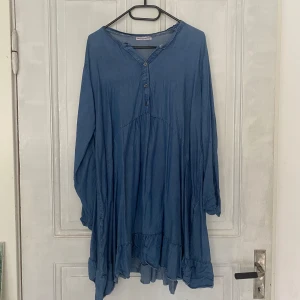 Blå långärmad klänning med volang från New Collection - Säljer en blå långärmad klänning från New Collection med knappar framtill och volangdetalj nedtill. Klänningen har en lös passform och är tillverkad i ett mjukt material som liknar jeans eller bomull. Perfekt för en avslappnad stil.