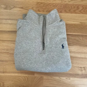 Grå halvzip tröja från Polo Ralph Lauren - Säljer en klassisk grå halvzip sweatshirt från Polo Ralph Lauren med broderad marinblå logga på bröstet. Tröjan har lång ärm, hög krage och ribbade muddar. Perfekt för en avslappnad och stilren look.