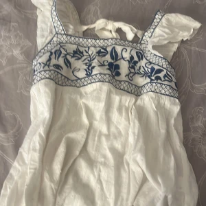 Vit klänning med blå broderier och knytband - Superfin vit klänning med blå broderier upptill och knytband i ryggen. Axelbandsmodell med volangdetaljer och lätt struktur i tyget. Perfekt för sommarens alla tillfällen!