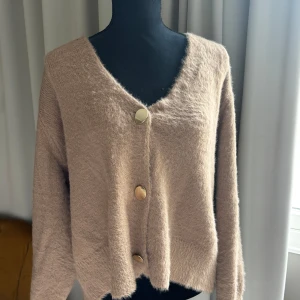 Beige fluffig kofta  - Mysig beige kofta från Primark med stora, ovala guldknappar framtill. Koftan är fluffig och har en avslappnad passform med långa ärmar och v-ringning. Perfekt för lager-på-lager under kyliga dagar.