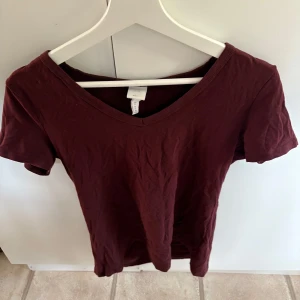 Vinröd t-shirt med v-ringning från H&M - En enkel vinröd t-shirt från H&M Basics med v-ringad hals och korta ärmar. Perfekt till vardags och lätt att matcha med olika outfits. Tillverkad i mjuk bomull för en bekväm känsla.