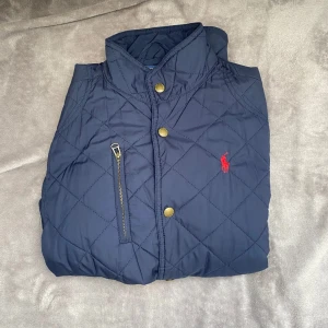 Mörkblå quiltad jacka från Ralph Lauren - Snygg mörkblå quiltad jacka från Ralph Lauren med guldiga knappar och dragkedja på bröstet. Jackan har ståkrage och det klassiska röda Polo-loggan broderad på bröstet. Perfekt för kyliga dagar.