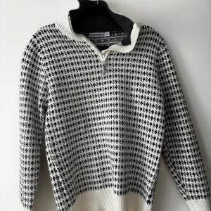 Vit och svart stickad tröja från Stormberg - Säljer en stickad tröja från Stormberg i vitt och svart med halv dragkedja och mönster över hela tröjan. Tröjan har hög krage och långa ärmar, perfekt för kyligare dagar.