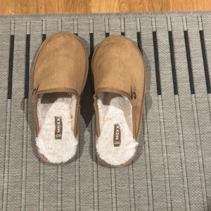 Bruna tofflor från Mexx - Mysiga bruna tofflor från Mexx med mjukt vitt foder och ovandel i mockaliknande material. Perfekta att glida runt i hemma, med en platt sula och enkel slip-in design. Säljes i storlek 42/43. Kontakta gärna vid frågor, mitt pris 85kr