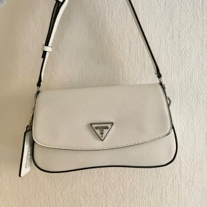Guess Cordelia axelväska - Säljer en stilren vit axelväska från Guess med silverfärgad logga framtill. Väskan har justerbart axelband och snygga metall-detaljer. Perfekt för att lyfta din outfit med en clean och modern touch. Finns små tecken av användning på axelremmen som inte är synliga vid användning💛