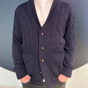 Mörkblå stickad kofta med knappar - Säljer en mörkblå stickad kofta med kabelstickat mönster och stora bruna knappar. Koftan har två fickor framtill och bred krage. Perfekt för lager-på-lager och passar till det mesta.