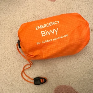 Orange nödbivack i syntetmaterial - Säljer en orange nödbivack för utomhusbruk, perfekt att ha med sig på äventyr eller camping. Kommer i en kompakt påse med dragsko och är tillverkad i lätt och slitstarkt syntetmaterial. Enkel att packa ner och ta med i väskan.Överlevnadssovsäck, Nöd Bivvy Bag PE aluminiumfilm Nödfilt Bushcraft för utomhuscamping och vandring Produkt Värmebevarande: Bivvy nödpåsar ger värmebevarande och termisk isolering i överlevnadssituationer. Fodrad med värme reflekterande material som liknar nödfiltar, detta