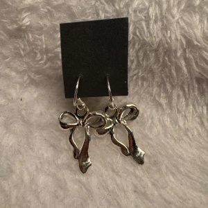 Silverfärgade örhängen med rosett - Säljer ett par snygga silverfärgade örhängen i form av rosetter. Perfekta för att ge din outfit en söt och trendig touch. Hängande modell som passar till många olika stilar. Krokarna är i sterling silver 