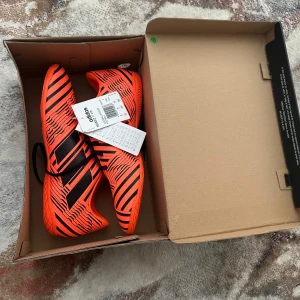 Adidas Nemeziz fotbollsskor i orange och svart - Säljer ett par Adidas fotbollsskor i en riktigt snygg orange färg med svarta zebraränder. Skorna har snörning och platt sula, perfekta för inomhusspel. Märket syns tydligt på både sula och plös. Skorna är lätta och smidiga för snabba rörelser på planen. Har inte använt skorna helt oanvänt och pris kan såklart diskuteras.