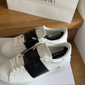 Vita sneakers Valentino med svart detalj - Säljer ett par vita sneakers från Valentino med en bred svart läderdetalj över vristen. Skorna har rund tå, platt sula och klassisk snörning. Perfekta för dig som gillar stilrena och lyxiga sneakers med en twist.