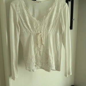 Vit Odd Molly blus med broderi - Supersöt vit blus från Odd Molly med v-ringning, broderade detaljer och knyt framtill. Långärmad och i mjuk bomull, perfekt för en romantisk och bohemisk look. 
