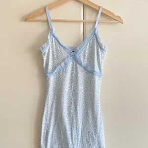 Blommig ljusblå kortklänning med spets - Söt kortklänning med ljusblå och vita små blommor, tunna axelband och spetsdetaljer längs halsringningen och under bysten. Klänningen har en omlott-v-ringad front och ärmlös design, perfekt för sommarens varma dagar.