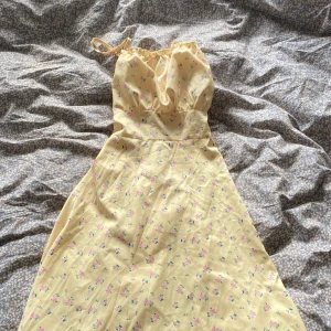 Gul medellång klänning med blommigt mönster - Säljer en söt gul medellång klänning med tunna axelband och blommigt mönster i rosa och grönt. Klänningen har snörning i ryggen och en lätt volang vid halsen. Perfekt för varma dagar och har en feminin, flowy siluett. Ger en otroligt snygg midja. 