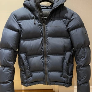 Mörkblå dunjacka Peak Performance Small - Säljer en mörkblå dunjacka från Peak Performance i storlek S. Jackan har en puffig look, huva, dragkedja framtill och två fickor. Perfekt för kalla dagar och riktigt snygg till streetwear-stilen.