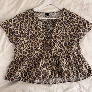 Leopardmönstrad blus från Gina Tricot - Säljer en superfin blus från Gina Tricot med leopardmönster i beige, brunt och svart. Toppen har korta ärmar, volang nertill och snörning framtill. Den är knappt använd. 