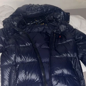 Mörkblå pufferjacka från Ralph Lauren - Säljer en glansig mörkblå pufferjacka från Ralph Lauren med huva och dragkedja. Jackan har en klassisk quiltad look och är perfekt för kalla dagar. Detaljerad med liten röd logga på bröstet och praktiska fickor. Snygg och stilren design som sticker ut.