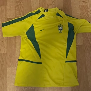 Brasilien Ronaldo 9 fotbollströja Nike - Gul Brasilien fotbollströja från Nike med gröna detaljer och tryck. På framsidan finns landslagets emblem och Nike-logga, på baksidan står det 'Ronaldo' och nummer 9. Tröjan är kortärmad och har sportig passform, tillverkad i lätt polyester.