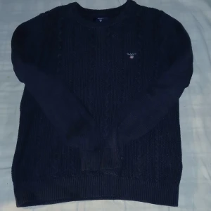 Mörkblå stickad tröja från GANT - Mörkblå stickad tröja från GANT med klassiskt kabelstickat mönster och rund hals. Tröjan har långa ärmar och ribbade muddar vid ärmslut och nederkant. Perfekt för kyliga dagar och snygg till jeans eller chinos.