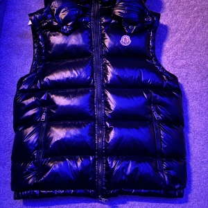 Svart dunväst från Moncler - Snygg svart dunväst från Moncler med glansig finish och klassisk logga på bröstet. Västen har dragkedja framtill, två fickor med dragkedja och hög krage. Perfekt för lager-på-lager och ger en cool look till din outfit.