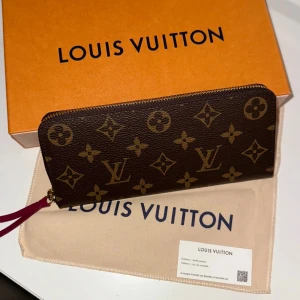 Louis Vuitton monogram plånbok - Snygg plånbok från Louis Vuitton i klassiskt monogrammönster. Utsidan är brun med guldiga LV-loggor och detaljer, insidan är vinröd med flera kortfack och dragkedja. Materialet är skinn och textil, perfekt för dig som gillar lyxiga accessoarer.
