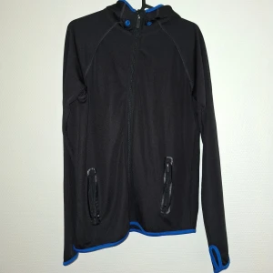 Svart sportig vindjacka från KappAhl - Svart vindjacka från KappAhl med blå detaljer och huva. Jackan har dragkedja framtill, två fickor med dragkedja och blå text 'MOOD SPORT' längs ryggen. Tillverkad i lätt polyester, perfekt för aktiva dagar. Långärmad med tumhål och sportig vibe.