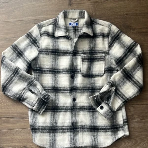 Rutig overshirt från Jack & Jones - Snygg overshirt från Jack & Jones i svartvitt rutigt mönster. Skjortan har klassisk krage, bröstficka och knäppning framtill. Tillverkad i mjukt material som känns lite ulligt, perfekt att slänga över en t-shirt. Långärmad och passar till lager-på-lager-stil.