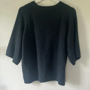 Svart stickad tröja med trekvartsärm - En svart stickad tröja med rund halsringning och trekvartsärmar. Tröjan har en enkel och stilren design med ribbade muddar vid hals, ärmslut och nederkant. Perfekt för lager-på-lager och chill dagar.