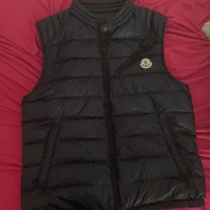 Svart dunväst från Moncler - Snygg svart dunväst från Moncler med klassisk quiltad design och hög krage. Västen har dragkedja framtill, två fickor och Moncler-logga på bröstet. Perfekt för lager-på-lager och ger en clean streetstyle-vibe. Den är äkta jag köpte den från NK