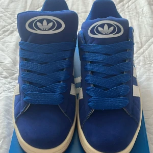 Blå Adidas Campus sneakers strl 42 - Snygga Adidas Campus sneakers i blå mocka med klassiska vita ränder på sidorna och vit sula. Skorna har rund tå, platt sula och blå snörning. Adidas logga på plösen och sidan. Perfekt för dig som gillar retrostil och streetwear.