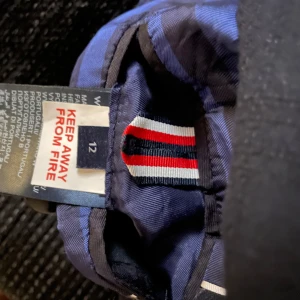 Mörkblå kavaj Tommy Hilfiger - Snygg mörkblå kavaj från Tommy Hilfiger med två knappar framtill och klassisk krage. Insidan har blått och svart randigt foder samt en innerficka med knapp. Detalj med röd, vit och marinblå rand på insidan av kragen. Perfekt för en stilren look.