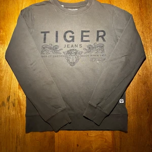 Grå sweatshirt från Tiger of Sweden Jeans - Snygg grå sweatshirt från Tiger of Sweden Jeans med stort svart tryck framtill med tigrar och logga. Tröjan har rund hals, långa ärmar och ribbade muddar. Perfekt för dig som gillar stilrena och coola plagg med streetkänsla. Storlek S och är liten i passformen. Någon fläck på enstaka ställen som går bort i tvätt. Vid funderingar är det bara att höra av sig jag är ärlig och rak med svar😁👍🏼