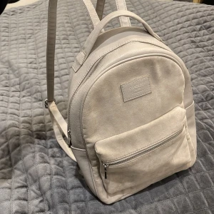 Beige väska - Beige ryggsäck med justerbara axelremmar och silvriga dragkedjor.      Mellanstor, ca 20 x 17 x 6 cm.                 Inga synliga defekter, endast lite smutsig inuti.