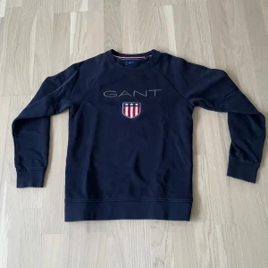 Mörkblå GANT sweatshirt med brodyr - Snygg mörkblå sweatshirt från GANT med broderad logga och sköld på bröstet. Tröjan har rund hals, långa ärmar och ribbade muddar. Klassisk och stilren design med röd, vit och blå detalj i nacken. Perfekt för dig som gillar preppy stil.