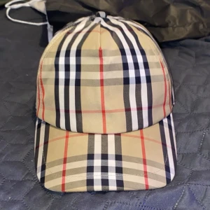 Burberry  keps - Snygg Burberry keps med klassiskt rutigt mönster i beige, svart, vitt och rött. Justerbar baktill med svart spänne och tydlig Burberry London England patch. Tillverkad i bomull och polyester, perfekt för dig som vill ha en ikonisk streetstil. Pris kan diskuteras!