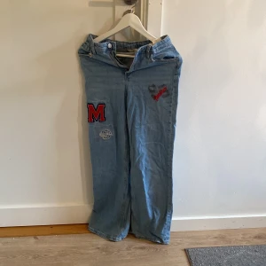 Blå jeans med broderade patches - Snygga ljusblå jeans med bred passform och coola broderade patches på framsidan, bland annat ett rött M, en New York-glob och Brooklyn-text. Jeansen har klassiska bakfickor och bälteshällor. Perfekt för dig som vill sticka ut med streetstyle.