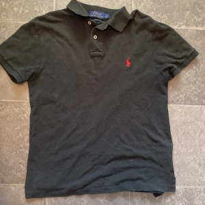Svart Polo Ralph Lauren piké tröja - Svart t-shirt från Polo Ralph Lauren i slim fit-modell. Klassisk pikékrage med två knappar och rött broderat logotyp på bröstet. Tillverkad i mjuk bomull, perfekt för en clean och stilren look.