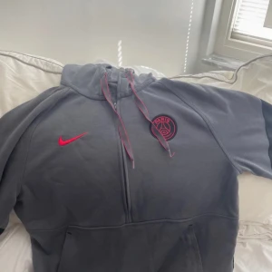 Grå PSG Nike hoodie med half zip - Säljer en grå Nike PSG hoodie med röd logga och snörning i huvan. Tröjan har half zip, Dri-FIT-material och en klassisk huva. Perfekt för dig som gillar sportig stil och vill representera Paris Saint-Germain.