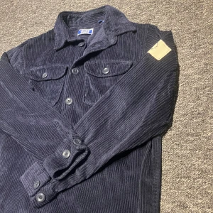 Mörkblå manchesterjacka Jack & Jones - Snygg mörkblå manchesterjacka från Jack & Jones med klassisk skjortkrage, två bröstfickor med knappar och en vit patch på ärmen (Ser lite beige ut men är vit i verkligheten). Jackan har raka linjer och stängs med knappar framtill. Perfekt för dig som gillar stilren och avslappnad look.