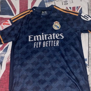 Real Madrid Vini Jr 21/22 - Snygg Real Madrid fotbollströja i marinblått med subtilt mönster, orange detaljer och tryck. Vini Jr. och nummer 7 på ryggen, klubbmärke och sponsor på bröstet. Kortärmad och tillverkad i lätt polyester som andas. Perfekt för dig som älskar fotboll och vill ha en cool look. Tröjan passar också i M