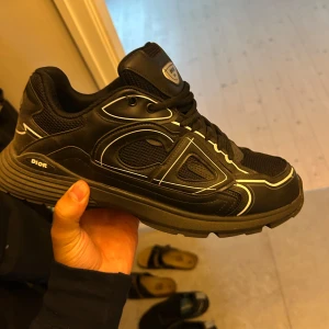 Svarta dior b30 - Säljer ett par svarta Dior sneakers med vita kontrastdetaljer och grov sula. Skorna har snörning, meshpaneler och coola grafiska linjer längs sidorna. Märket syns tydligt på plösen och innersulan. Perfekta för dig som gillar lyxig streetstyle.