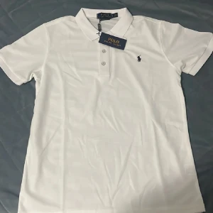 Vit Polo Ralph Lauren t-shirt🔥 - Vit t-shirt från Polo Ralph Lauren med klassisk pikékrage och korta ärmar. Slim fit-modell med tre knappar framtill och den ikoniska broderade loggan på bröstet. Tillverkad i mjuk bomull som känns skön mot huden. Perfekt för en clean och stilren look.